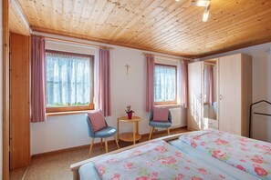 A room with a wooden ceiling, two windows with curtains, a bed with floral bedding, a small table with a plant, and a wardrobe. - Apartment 'Gisela' with Mountain View, Shared Terrace and Wi-Fi (Bernau im Schwarzwald)