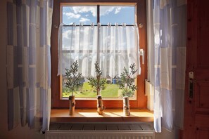 Ein Fenster mit durchsichtigen Vorhängen, drei kleine Blumentöpfe auf der Fensterbank und Blick ins Grüne draußen.