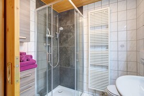 Ein modernes Badezimmer mit einer gläsernen Duschkabine, weißen Fliesenwänden, einer Holzbalkendecke und einem WC mit weißem Sitz.