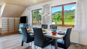 A dining area with a table set for four, a television, and large windows with a view of greenery.