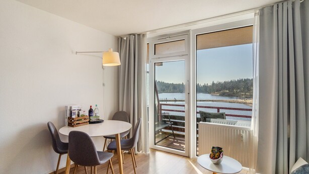 Ein Zimmer mit Esstisch, Stühlen, einem kleinen runden Tisch mit Schüssel, einer Lampe und Blick auf ein Gewässer durch große Fenster.