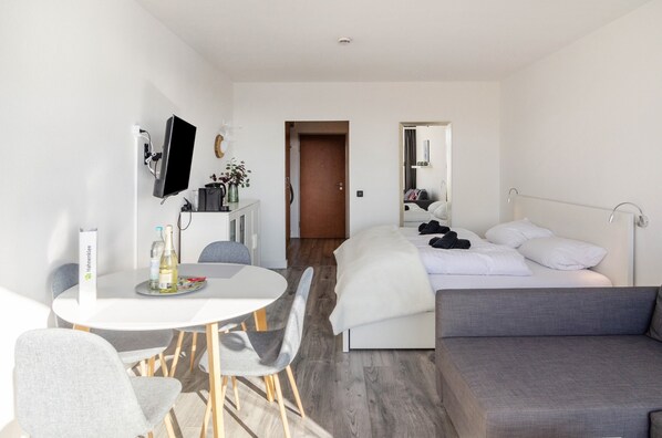 A modern hotel room with a bed, a sofa, a dining table with chairs, a TV, and a small desk with a computer and a bottle.