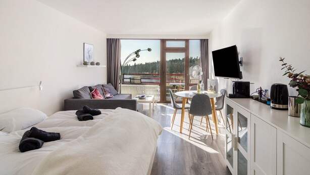 A modern hotel room with a bed, a sofa, a dining area with chairs and a table, a TV, and a balcony with a view of trees.