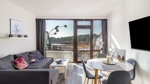 A modern living room with a sofa, coffee table, dining table, and chairs. There is a large window with a view of trees and a distant structure.