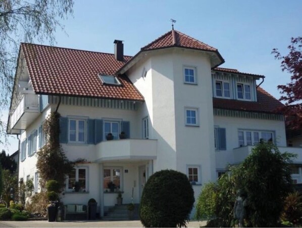 A two-story house with a red-tiled roof, a central chimney, and a symmetrical facade with multiple windows. - Apartment 'Häuschen' with Private Terrace, Shared Garden and Wi-Fi (Wasserburg (Bodensee))