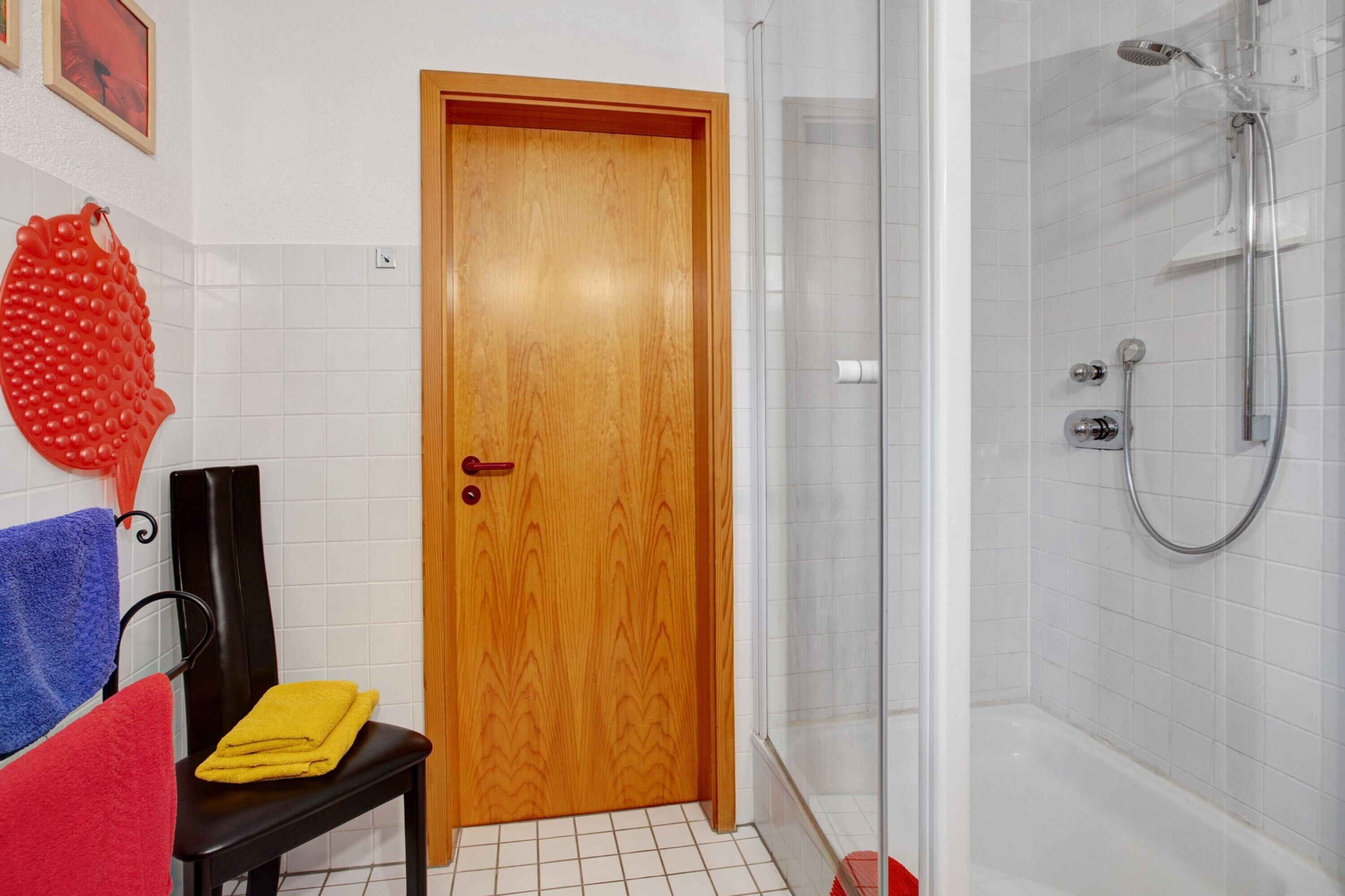 A bathroom with a wooden door, a shower with a glass enclosure, a chair with a yellow towel, and a red towel on the floor.