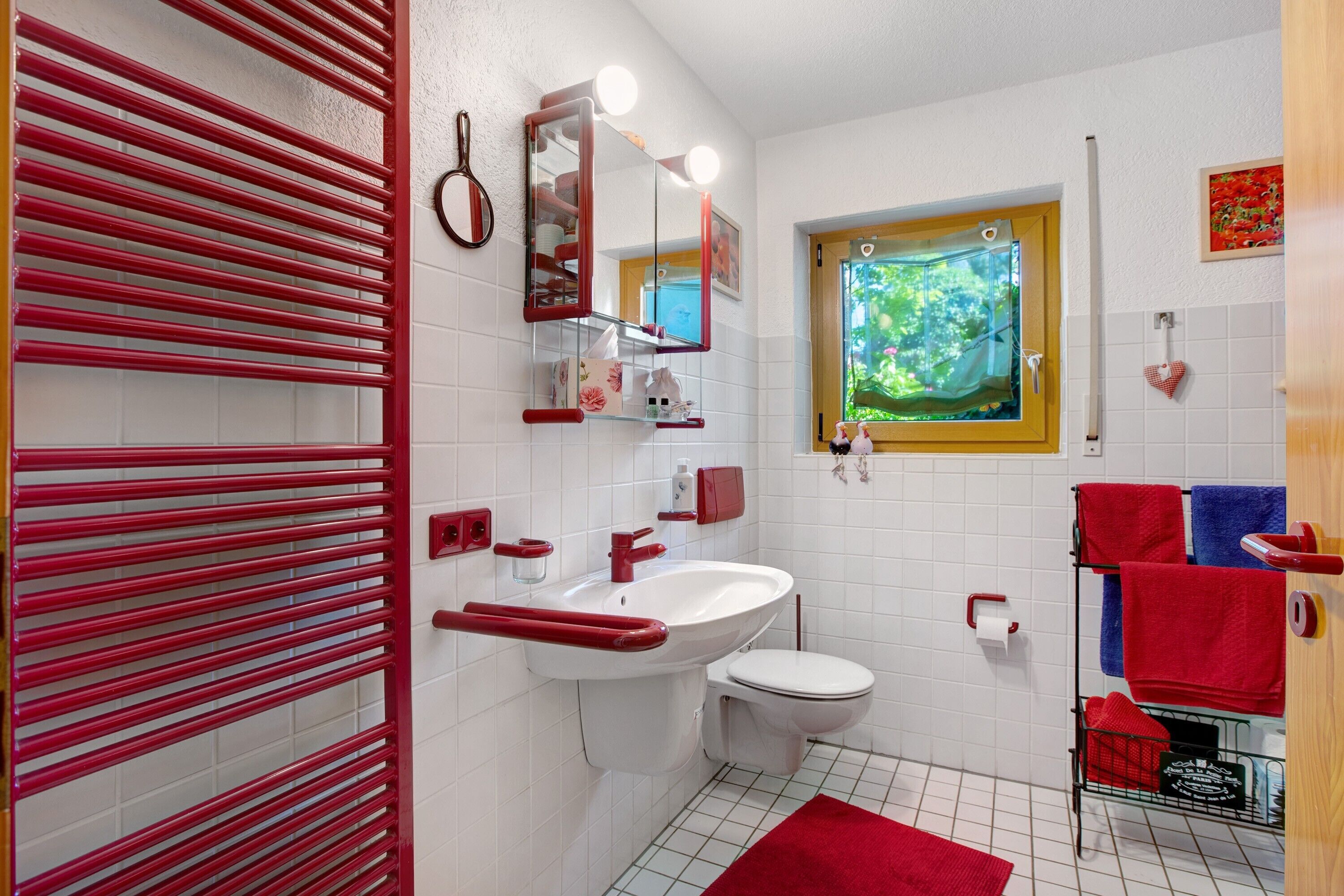 Ein Badezimmer mit einem weißen Waschbecken, einer Toilette und einem roten Handtuchhalter. Ein Fenster bietet Blick auf Grünflächen, und über dem Waschbecken befindet sich ein Spiegel mit Beleuchtung.