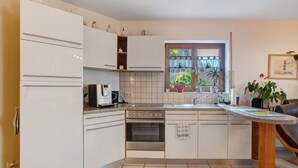 A compact kitchen with white cabinets, a built-in oven, a coffee maker, and a window with a view of greenery. - Apartment 'Nussbäumle' with Private Terrace, Shared Garden and Wi-Fi (Rheinfelden (Baden))