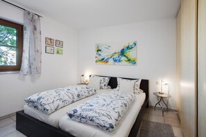 Un lit double avec une literie à motifs floraux, une table de chevet avec une lampe, un tableau abstrait accroché au mur et une fenêtre avec des rideaux.