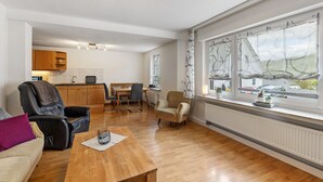 Een moderne woonkamer met een houten salontafel, een leren bank en fauteuils. Op de achtergrond is een eethoek met een tafel en stoelen zichtbaar. Grote ramen met behang gordijnen zorgen voor natuurlijk licht en uitzicht.