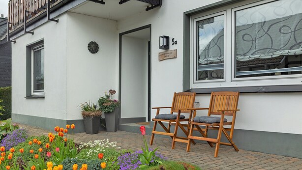 Een gebouw met een witte gevel, een deur en een raam met gordijn. Buiten staan twee houten stoelen.