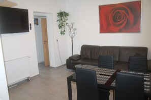 A living room with a sofa, a dining table with chairs, a TV mounted on the wall, and a large red rose painting.