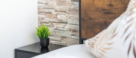 Una cama con cabecera, una mesita de noche con planta en maceta y una pared de piedra.