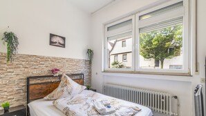 Quarto com parede de pedra, cama com roupa de cama estampada em folha, radiador e janela com vista para uma árvore e edifícios.