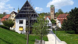Un edificio moderno con techo triangular, junto a casas tradicionales y una torre de iglesia.