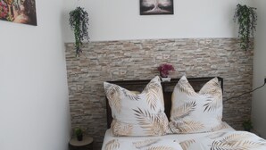 Un dormitorio con cabecero de pared de piedra, cama con ropa de cama estampada con motivos de hojas, una mesita redonda con una planta y un cuadro colgado en la pared.