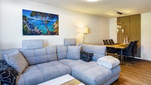 A modern living room with a gray sofa, a dining table with chairs, a painting of a coastal scene, and a ceiling light fixture.