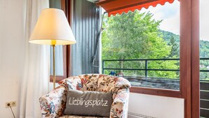 Un fauteuil à motifs floraux, agrémenté d’un coussin sur lequel est inscrit le mot « Lieblingsplatz », se trouve dans une pièce dont la fenêtre donne sur une zone boisée.
