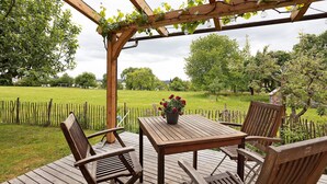 An outdoor wooden deck with a table and chairs, a pergola with climbing plants, and a view of a green field and trees.