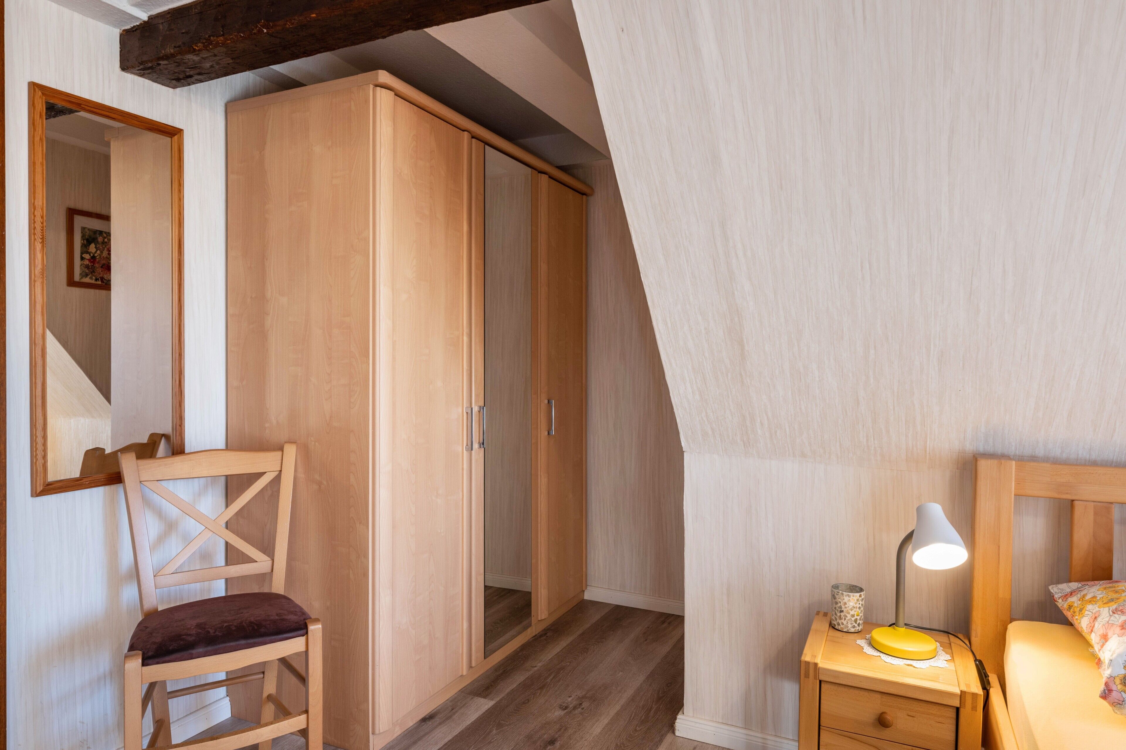 Une chambre à coucher avec une armoire en bois, une chaise, une table de chevet avec une lampe et un lit agrémenté d’un coussin à motifs floraux.