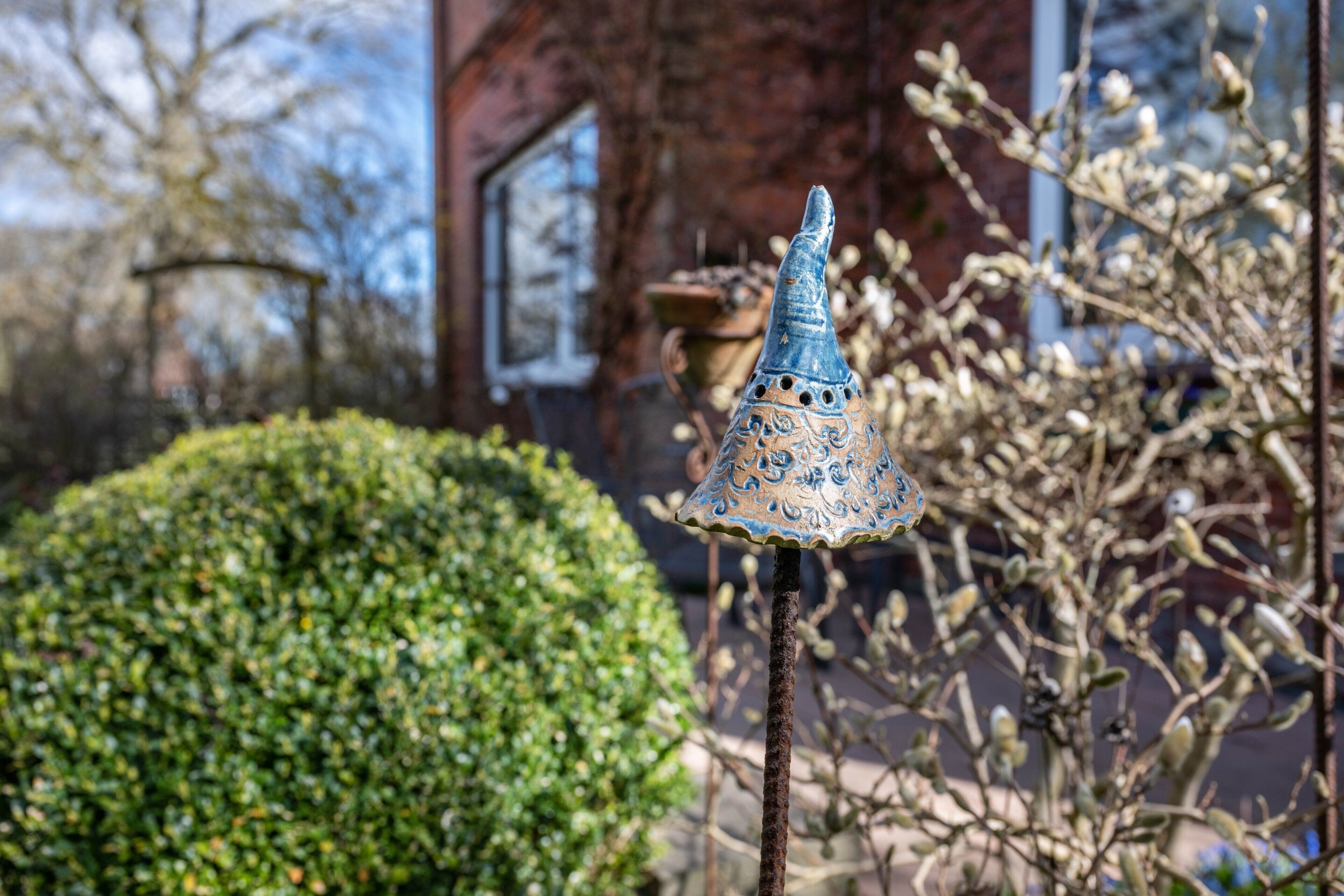 Un objet décoratif en métal, surmonté d’une partie conique, qui pourrait être un carillon éolien, placé dans un jardin avec une haie et un bâtiment en arrière-plan.