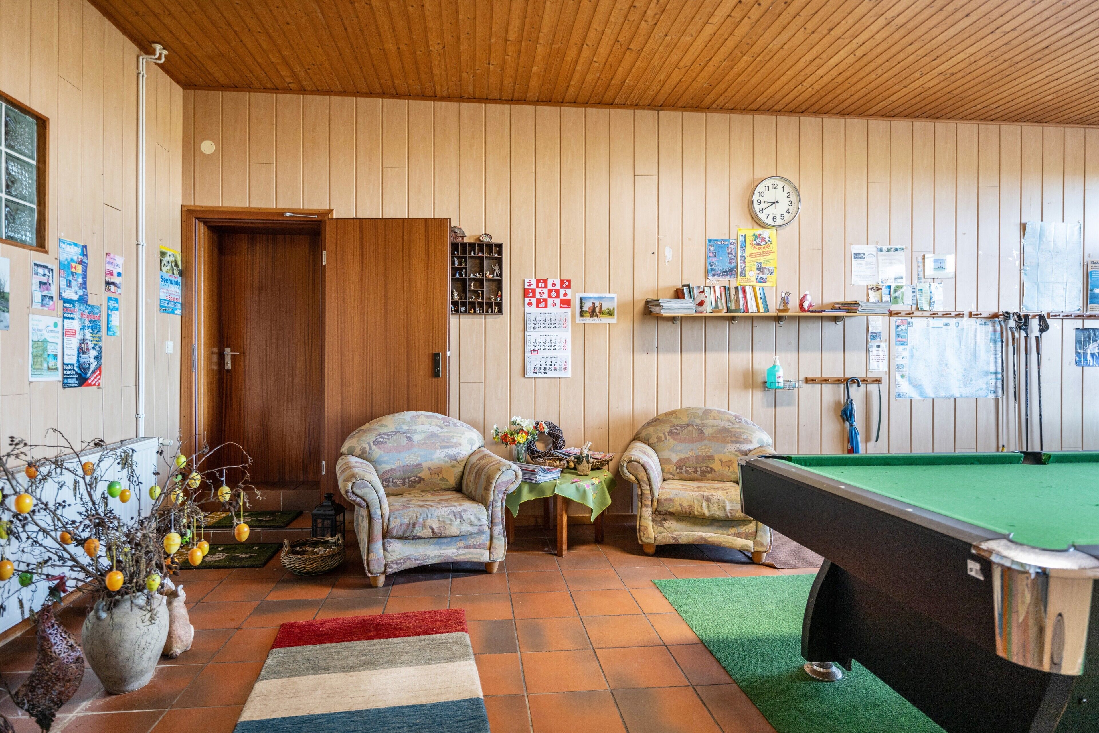 A room with wooden walls, a pool table, two armchairs, a clock, and various posters.