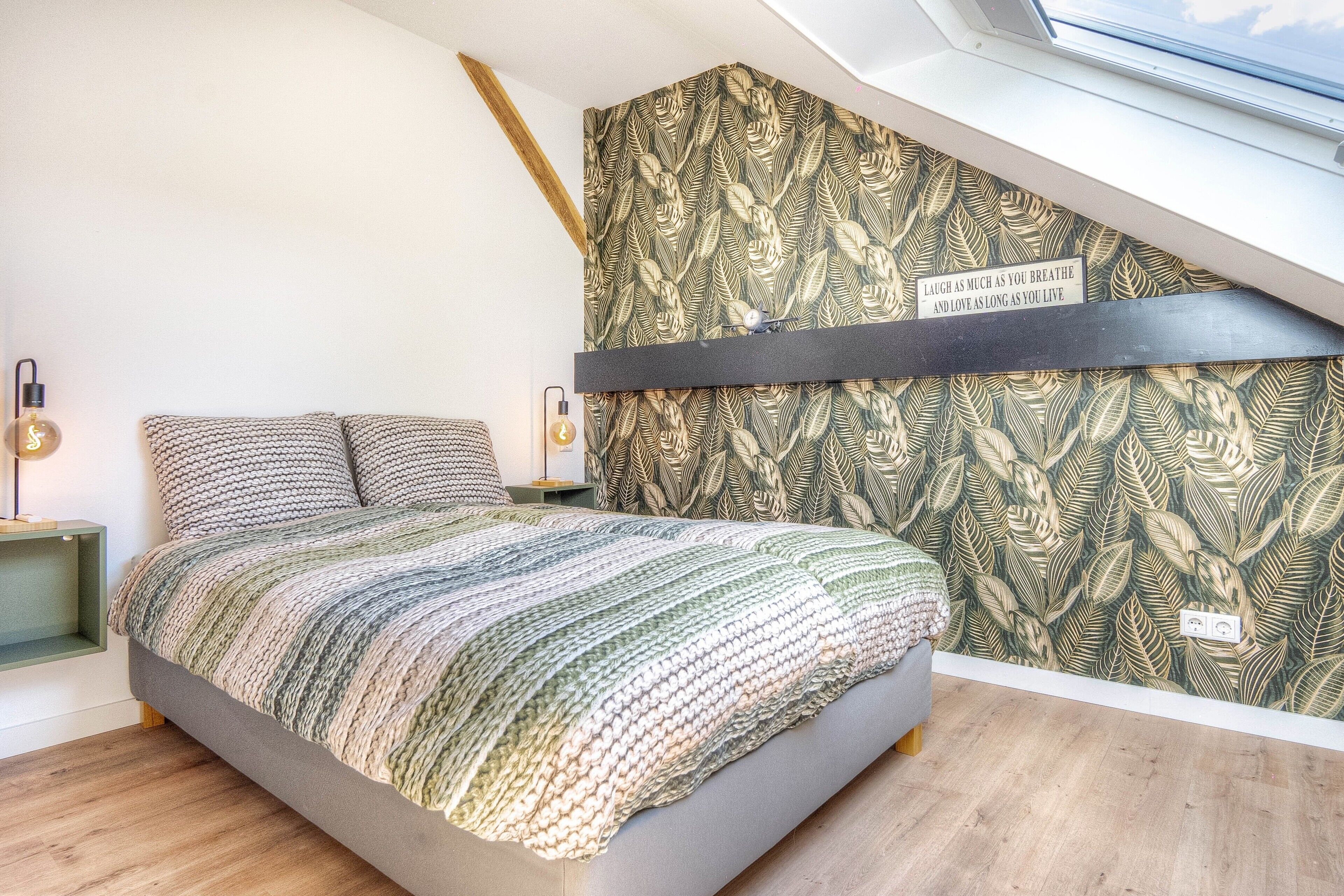 Ein Schlafzimmer mit schrägem Dach, einem Bett mit gestreifter Decke, einer Wand mit Blattmuster und einem Oberlicht.