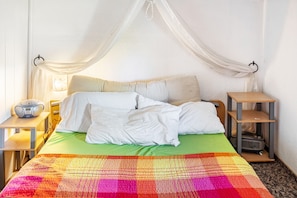 Un lit avec du linge de lit blanc, une couverture colorée, une table de chevet avec une radio et une petite table de nuit avec une lampe.