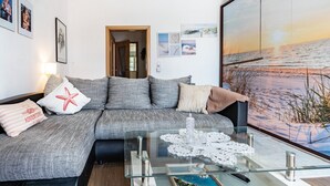Ein modernes Wohnzimmer mit einem grauen Sofa, einem Glastisch und einem großen Wandbild mit einer Strandansicht.