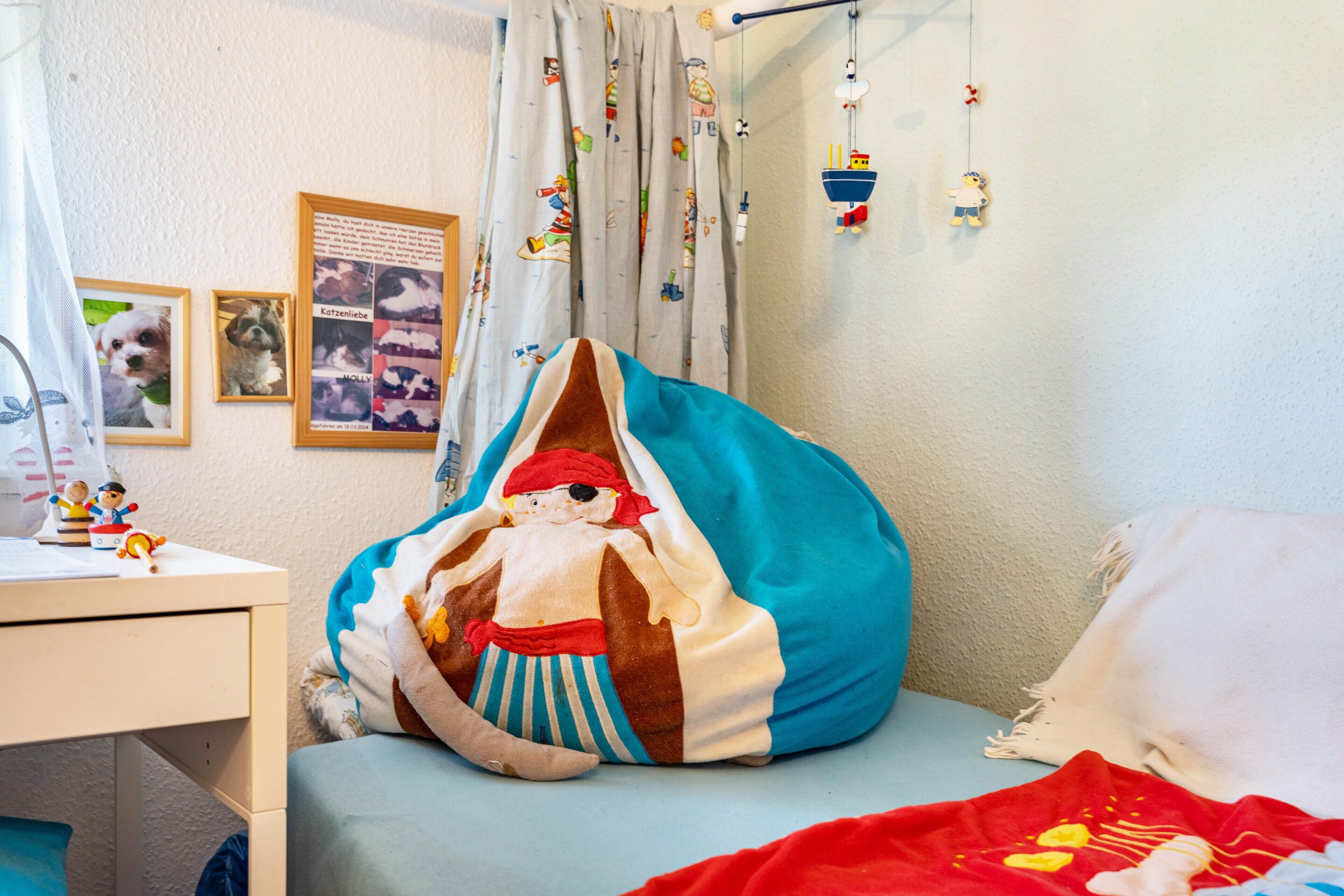 A child's bedroom with a bed, a desk, a chair, and a wall with framed pictures and a curtain.