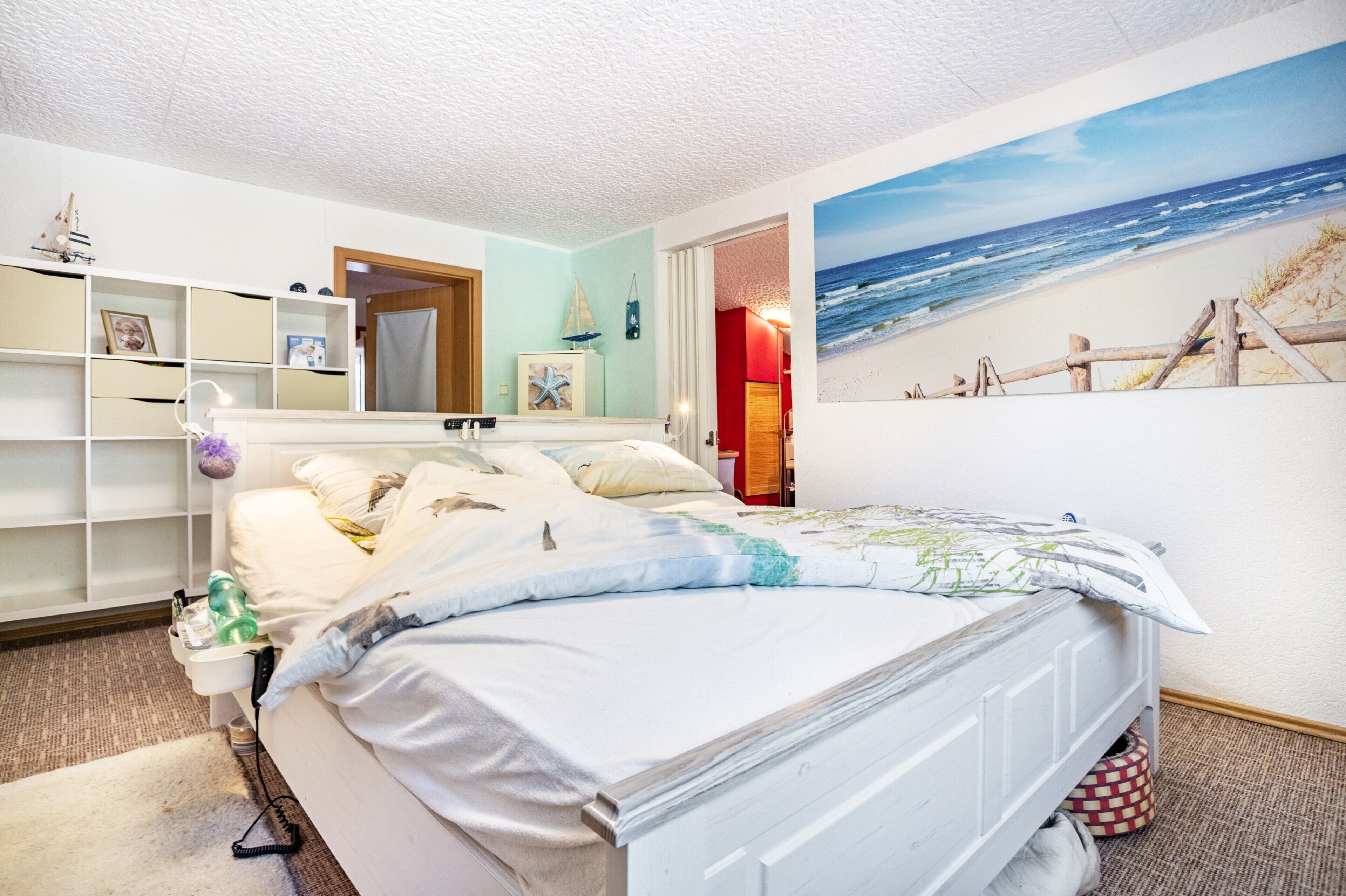 Un dormitorio con cama, una estantería, un estante en la pared, un cuadro de una playa y una puerta que da a otra habitación.
