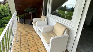 Une véranda couverte avec un sol carrelé, des meubles en osier peints en blanc et une vue sur la verdure.