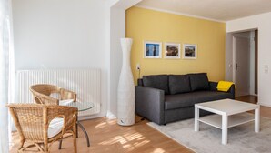 A living room with a sofa, two wicker chairs, a white coffee table, a tall white vase, and three framed pictures on the wall.