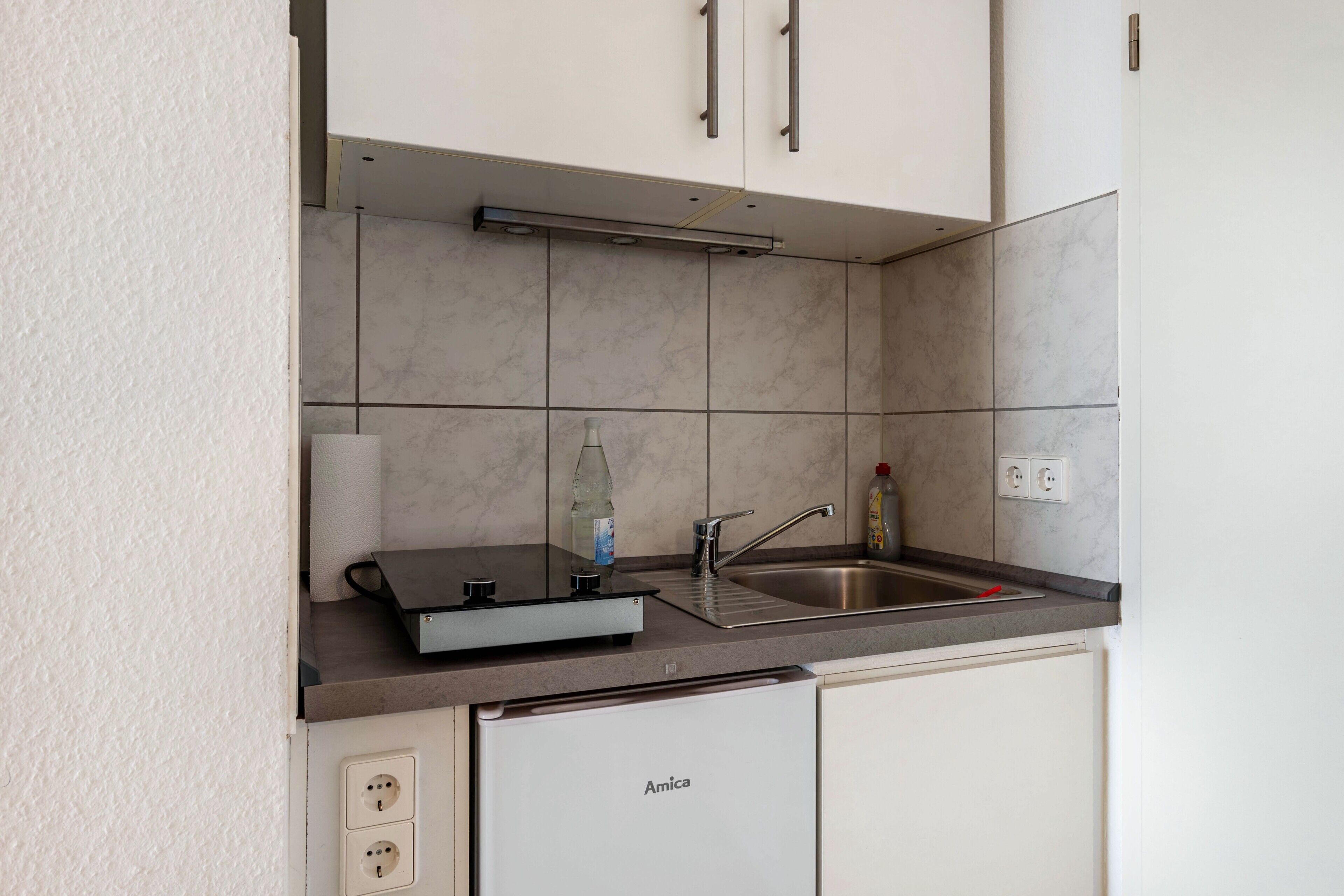 A compact kitchen with a built-in dishwasher, a sink, and a countertop with a bottle and a glass.