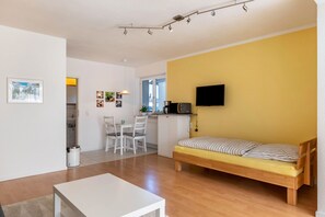 Un espace de vie compact comprenant un lit, une table à manger, des chaises, un coin cuisine et un téléviseur fixé au mur.