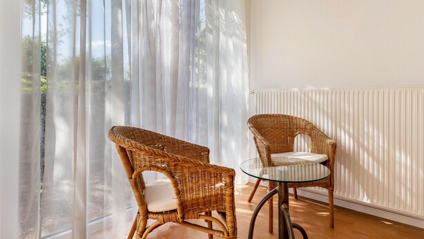 A room with two wicker chairs and a glass table, large windows with sheer curtains, and a radiator on the wall.
