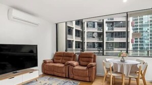 Un salon moderne doté d’un téléviseur à écran plat, de deux canapés en cuir marron, d’une table ronde en bois avec des chaises et d’une grande fenêtre offrant une vue sur la ville.