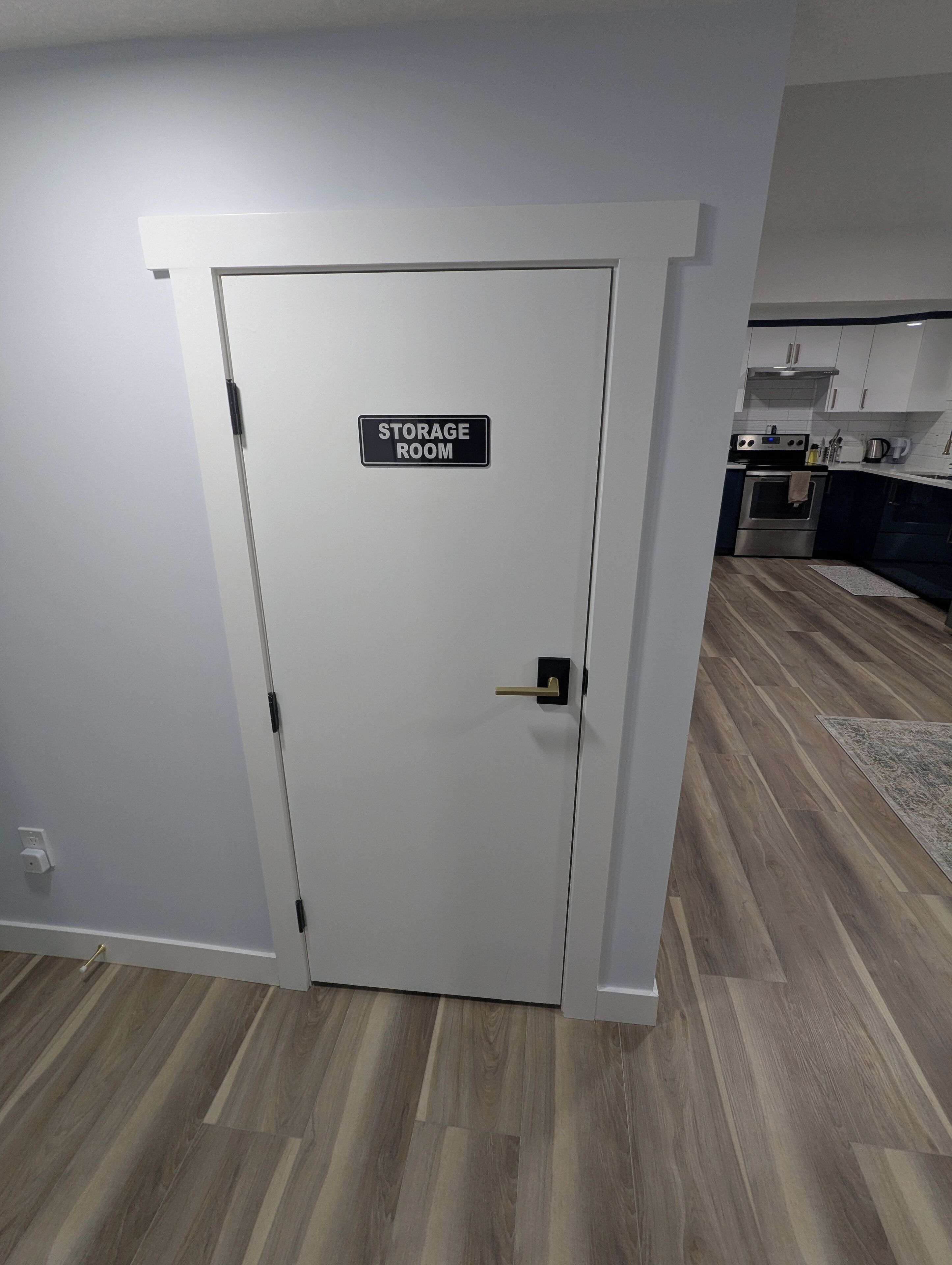 Une pièce de rangement avec une porte fermée, un coin cuisine avec des placards et des appareils électroménagers, et un plancher en bois.