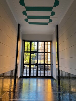 Un couloir doté d’une grande porte vitrée donnant sur l’extérieur, d’un sol en marbre et d’un plafond orné de motifs géométriques.