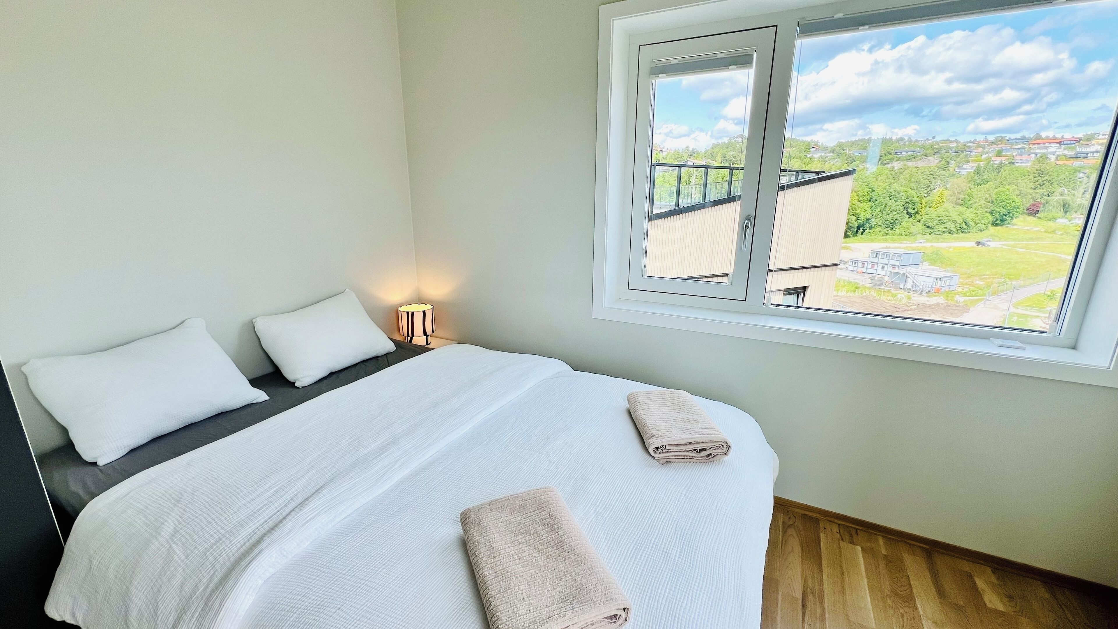 Een slaapkamer met een groot bed, twee kussens, een bedlamp en een raam met uitzicht op gebouwen en groen.