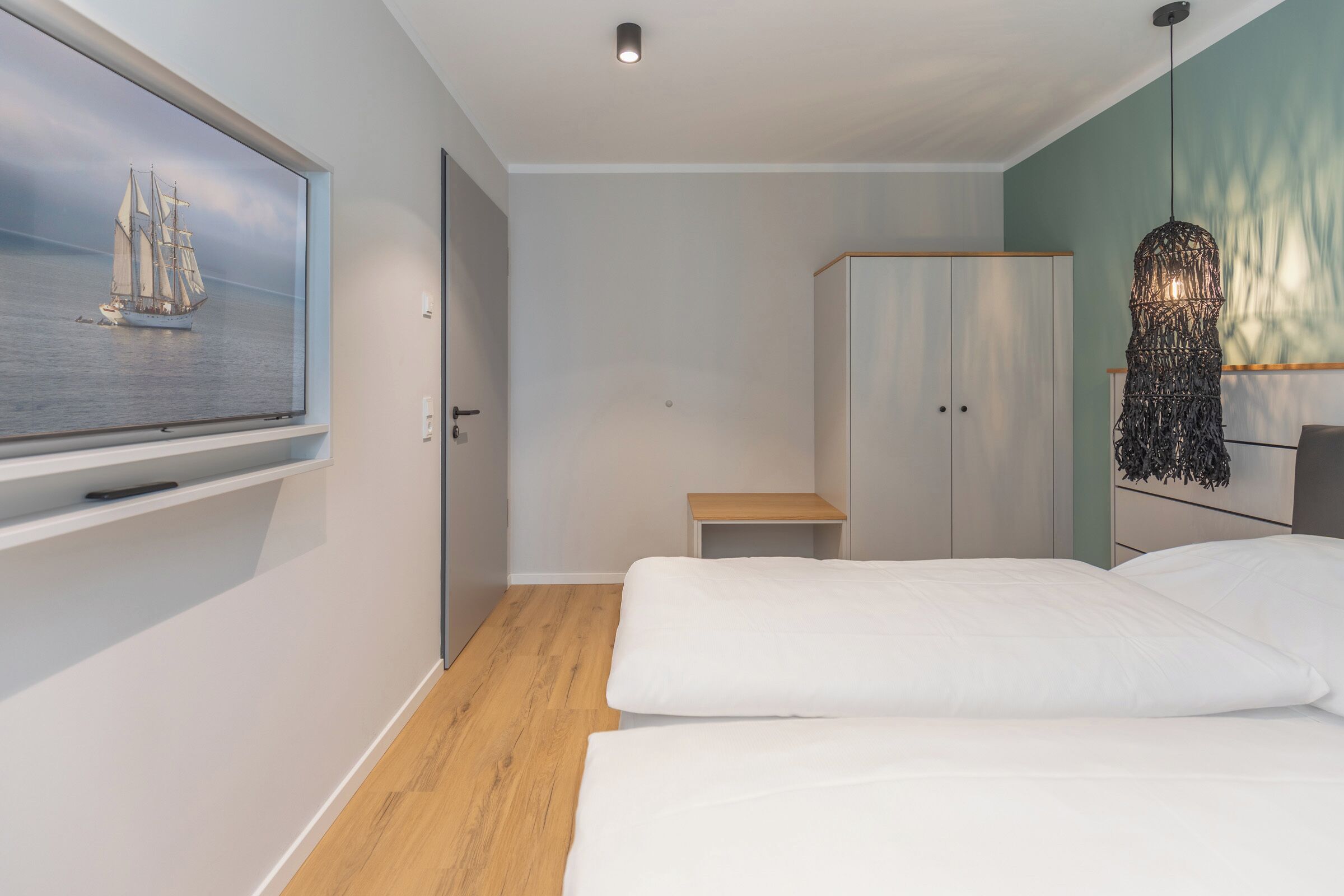 Ein modernes Schlafzimmer mit Bett, Holzboden, Kleiderschrank, einem kleinen Tisch und einem an der Wand montierten Fernseher, der ein Schiff auf dem Meer zeigt.