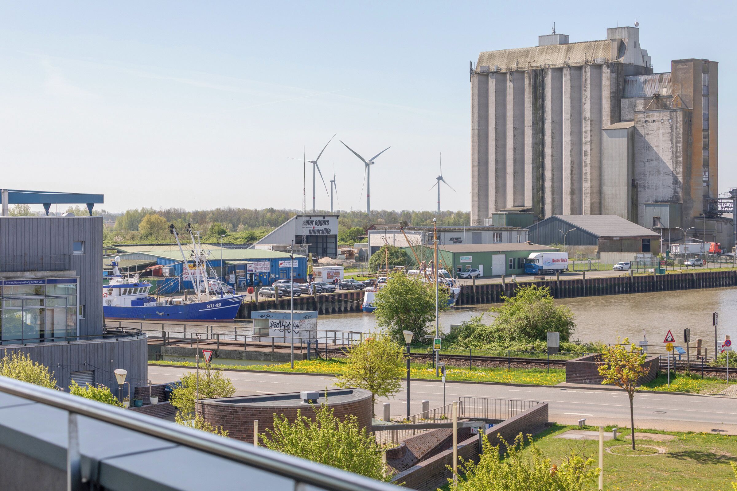Een rivierengebied met industrie, pakhuizen, een kade met boten en windturbines op de achtergrond.
