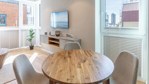 A modern living room with a wooden dining table, comfortable seating, a flat-screen TV mounted on the wall, and a large window offering a city view. - Apartment Zollkoje 03 directly at the Husum harbor (Husum)