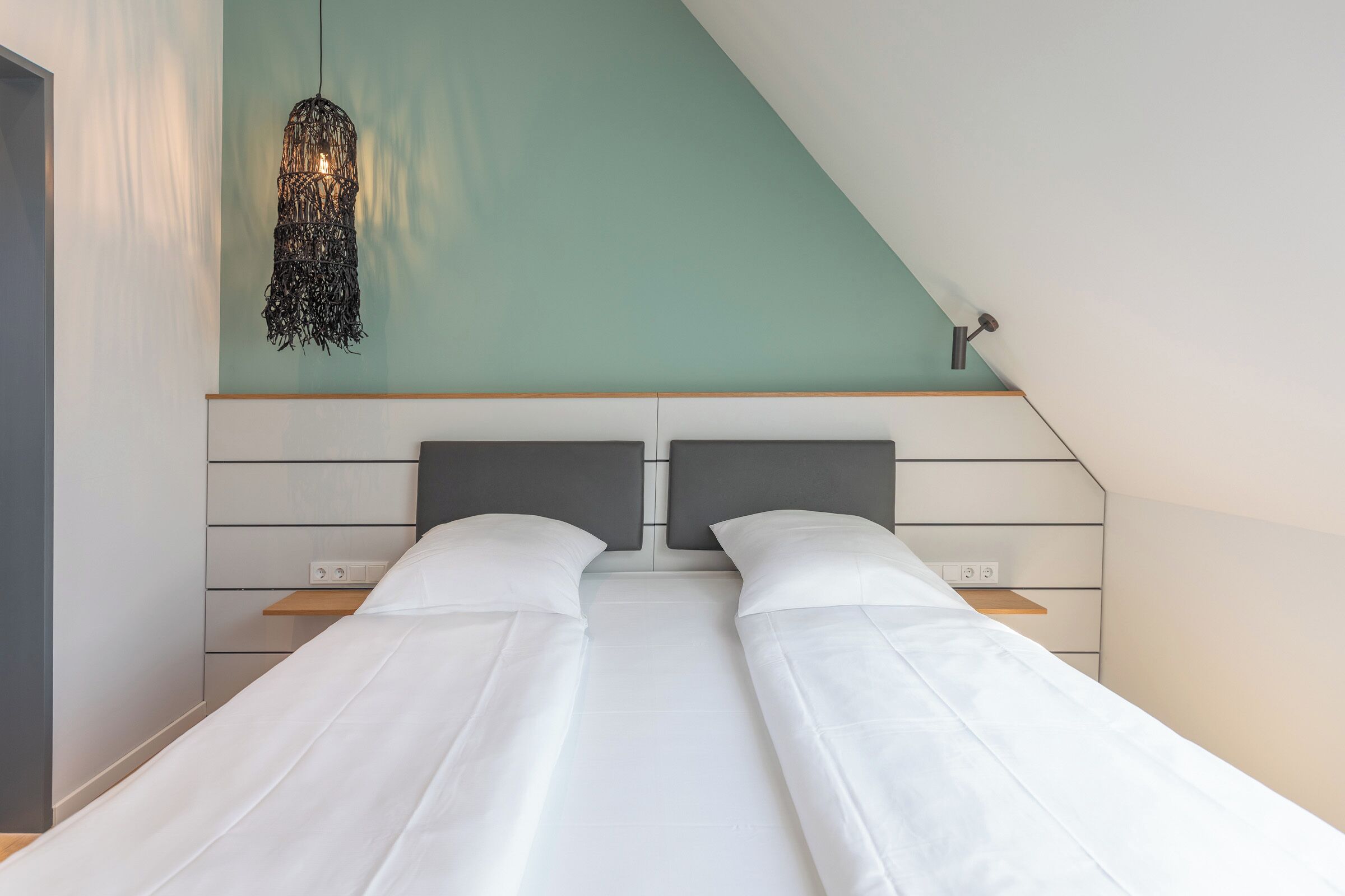 A modern bedroom with a sloped ceiling, two beds with white linens, a woven light fixture, and a wooden shelf.