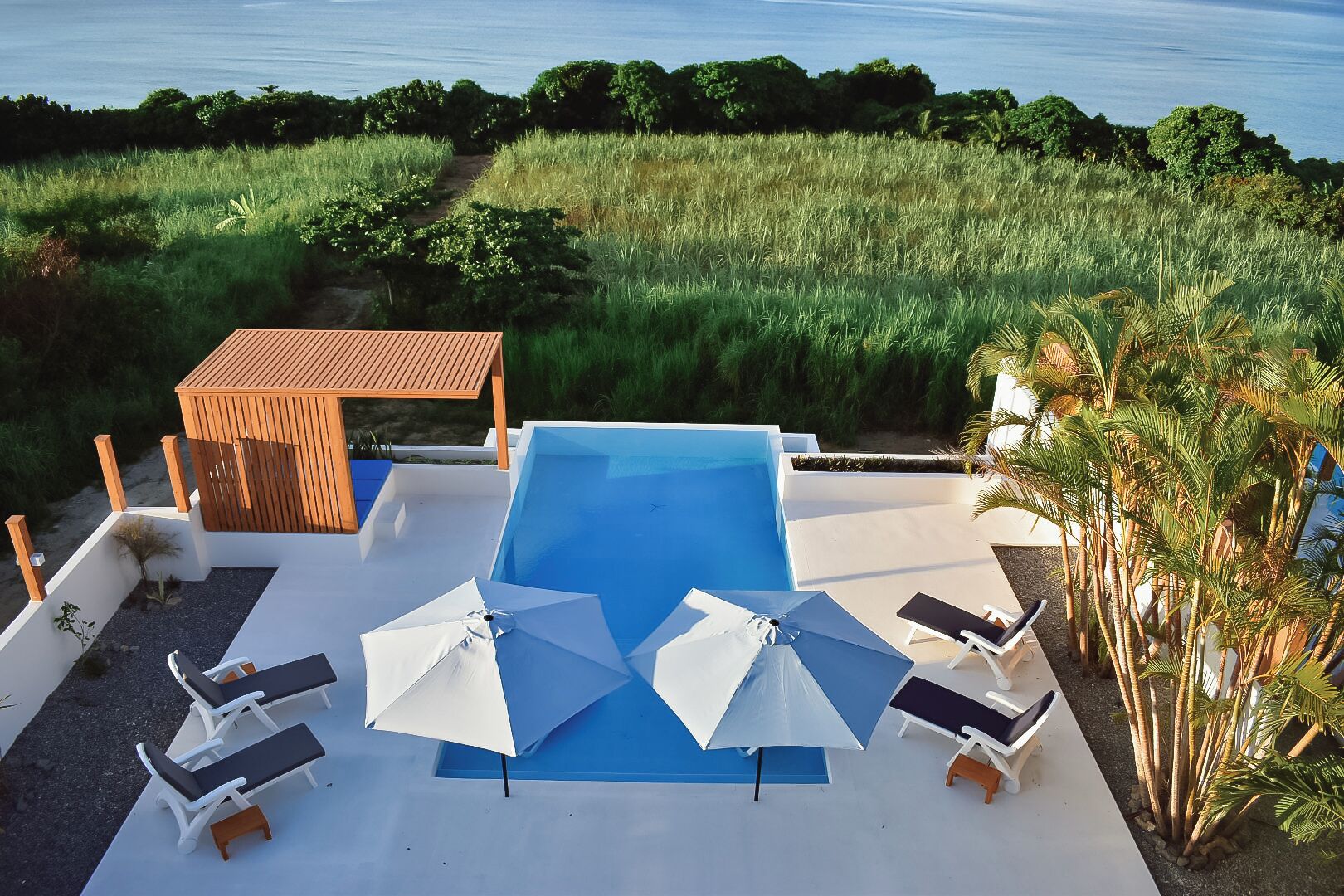 Zona de piscina con sillas de descanso y sombrillas, rodeada por una terraza de madera y vista al mar.