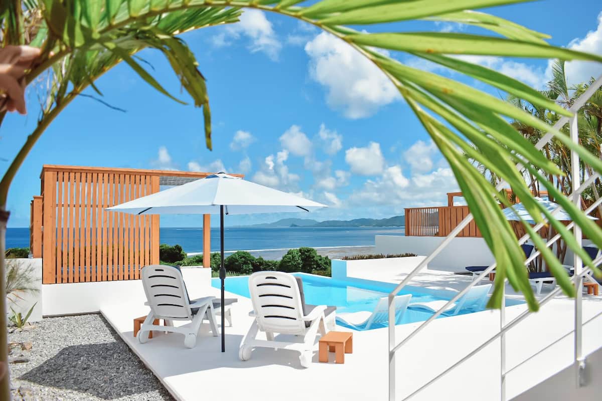 Ein Poolbereich mit zwei Liegestühlen und einem großen Sonnenschirm mit Blick auf Strand und Meer.