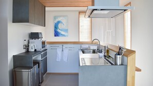 A modern kitchen with a built-in oven, microwave, and coffee maker. There is a sink with a faucet and a window with blinds. A painting of a wave is on the wall. - Ocean View Ishigaki Kaze & Nami (Ishigaki)