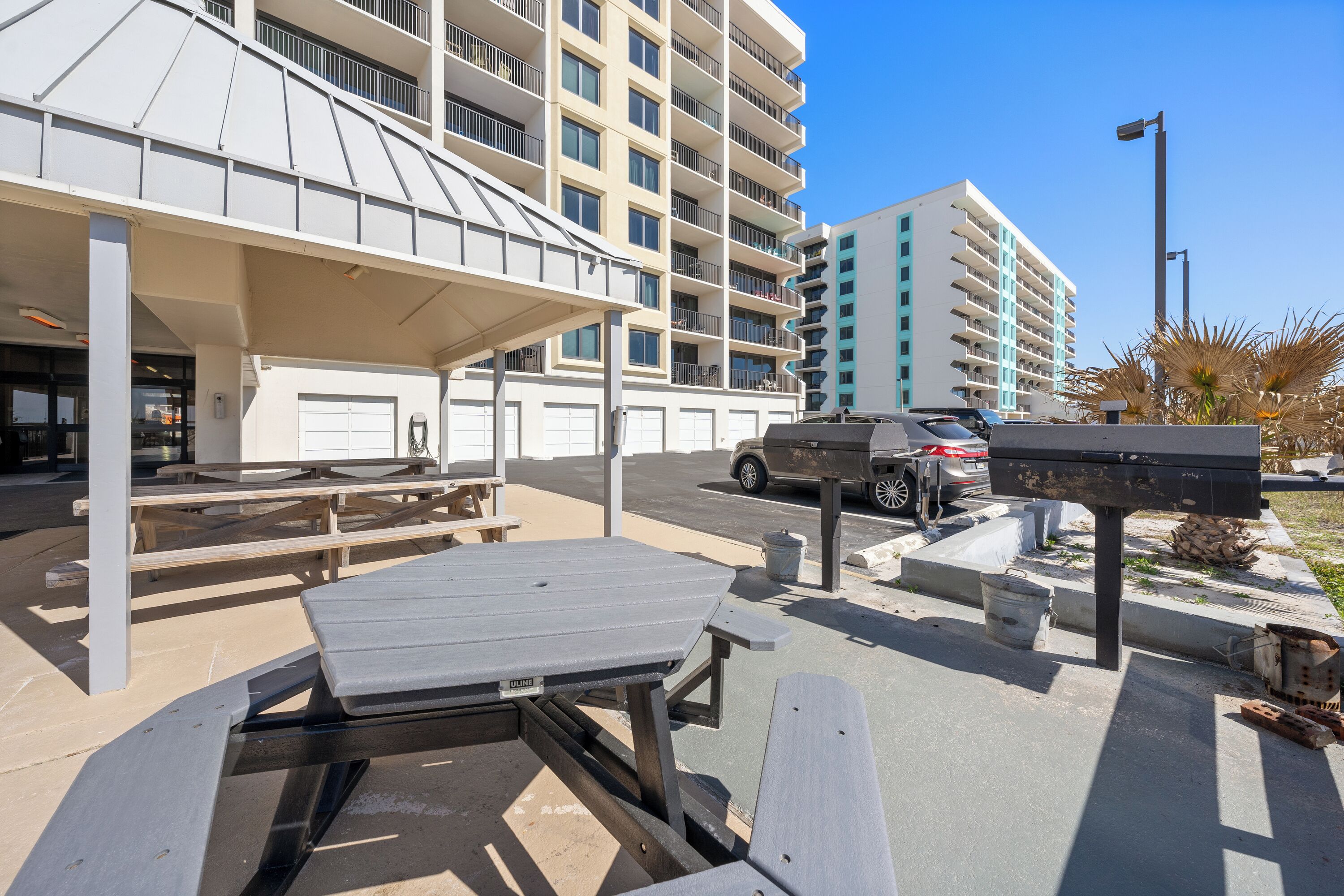 A parking area with picnic tables, a covered seating area, and a building with multiple balconies in the background.