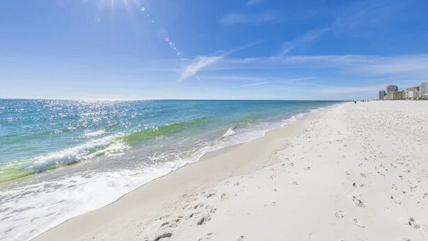 A beach with clear skies and a bright sun, featuring a sandy shore and a few buildings in the background. - Island Winds West 471 (Gulf Shores)