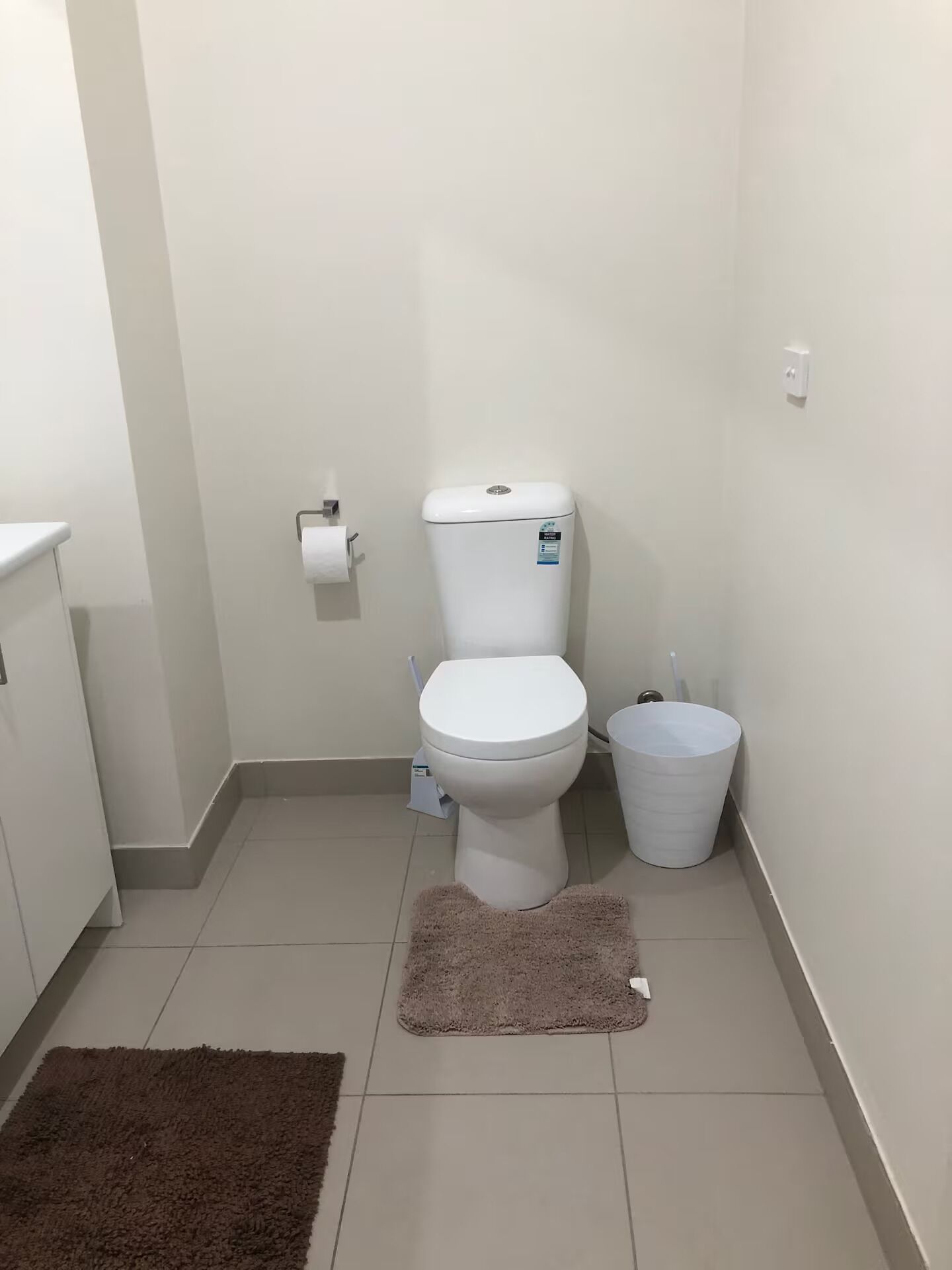 A small, clean bathroom with a toilet, a trash bin, and a single roll of toilet paper on the wall.